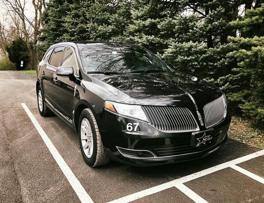 A black car is parked in a parking lot with trees in the background.