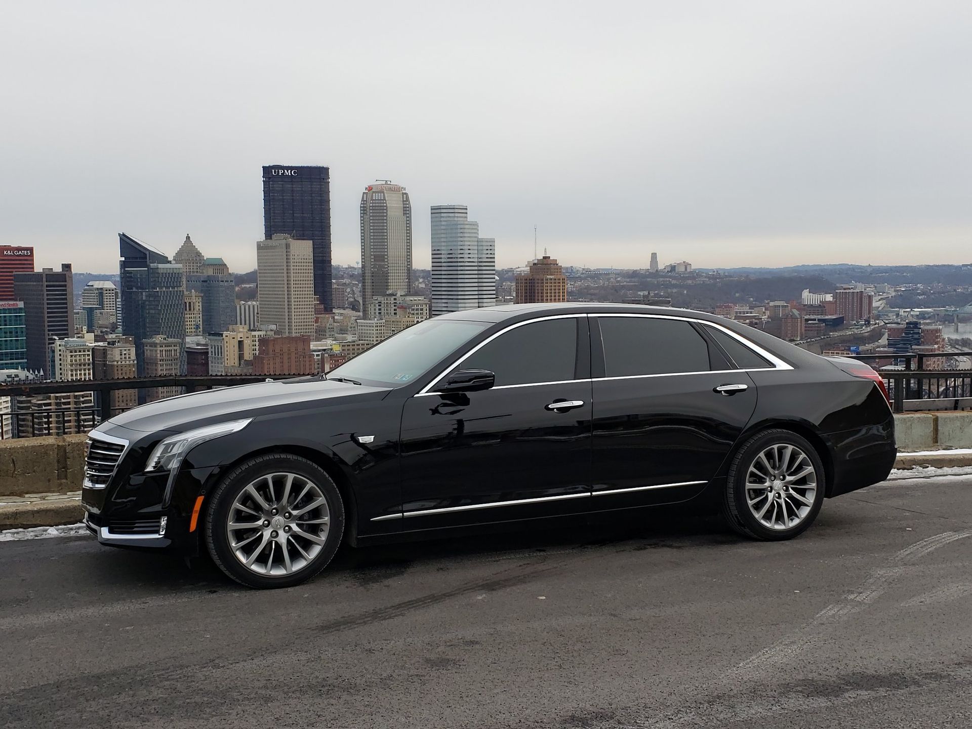 A black car is parked on the side of the road in front of a city skyline.