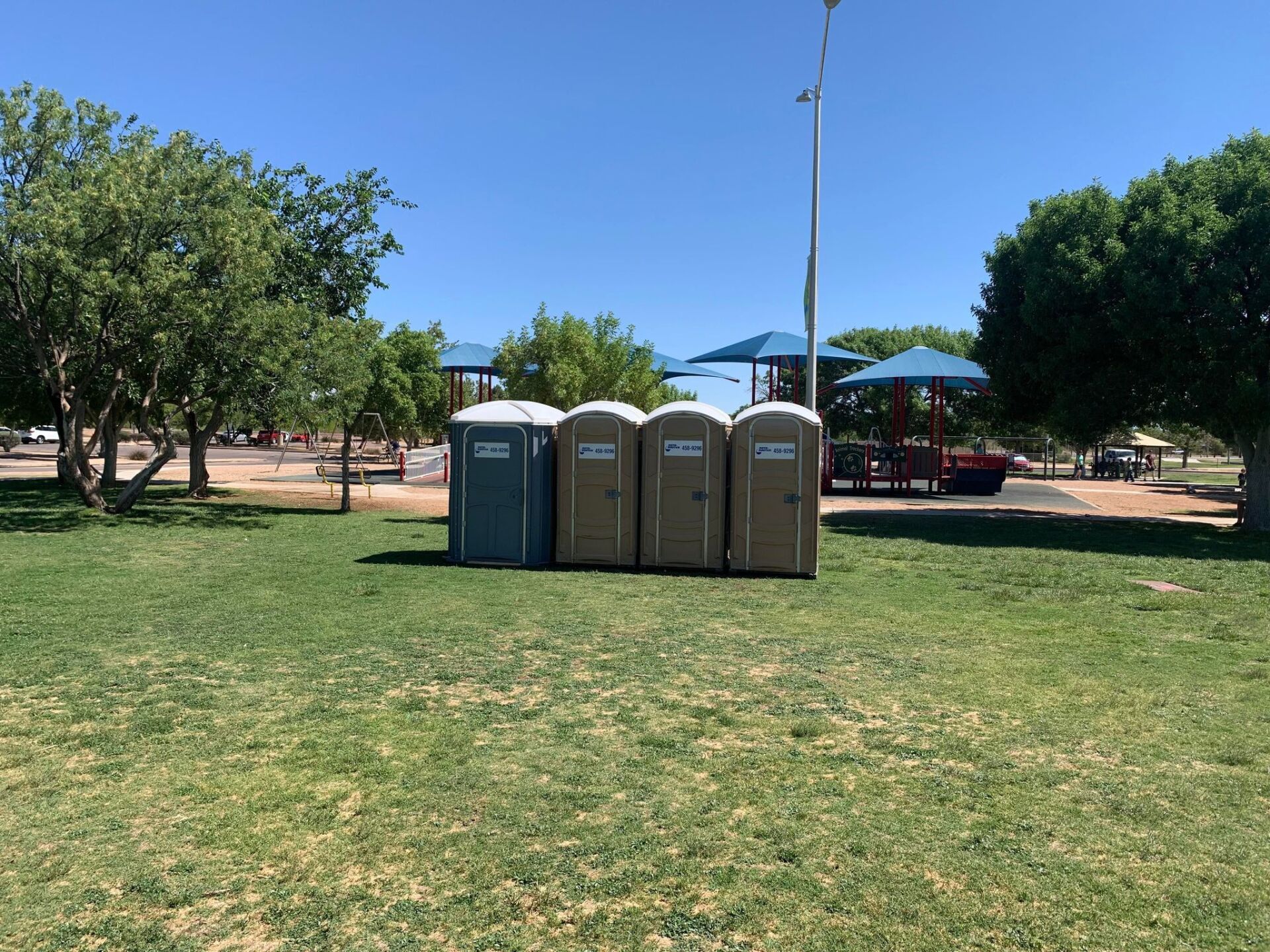 A row of portable toilets in a park with a playground in the background.