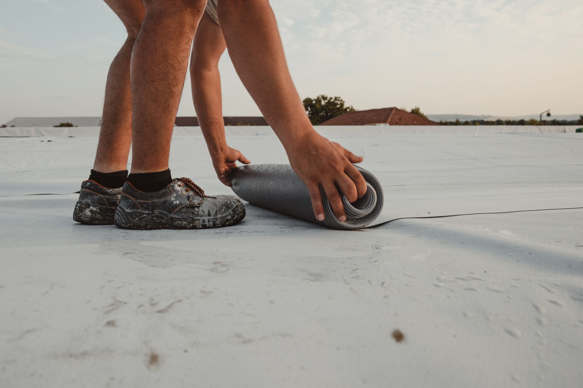 Person unrolling a gray roofing membrane on a white rooftop under a pale sky.