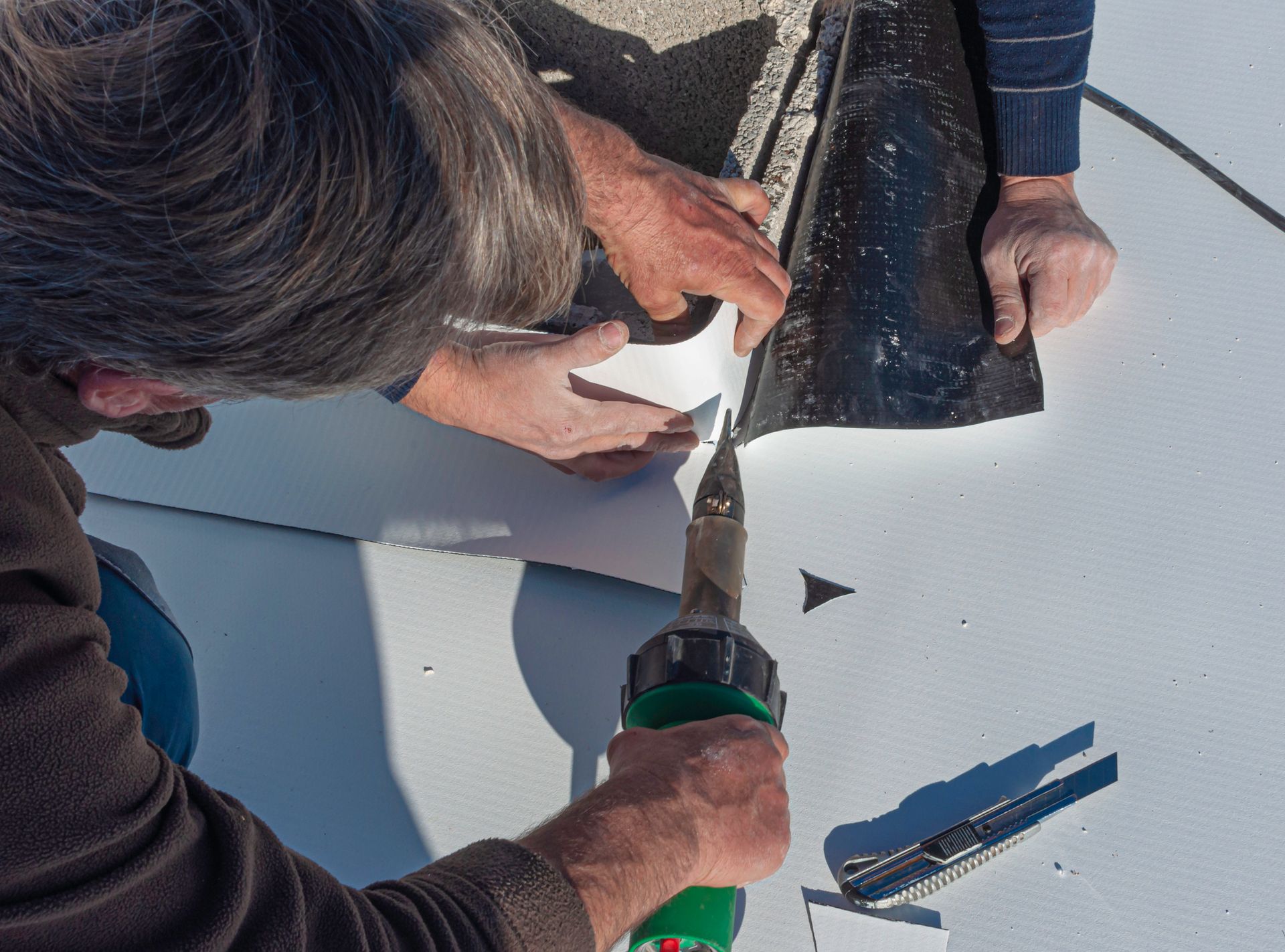 Two people working on a white surface, using a heat gun to weld a black sheet and white material together.