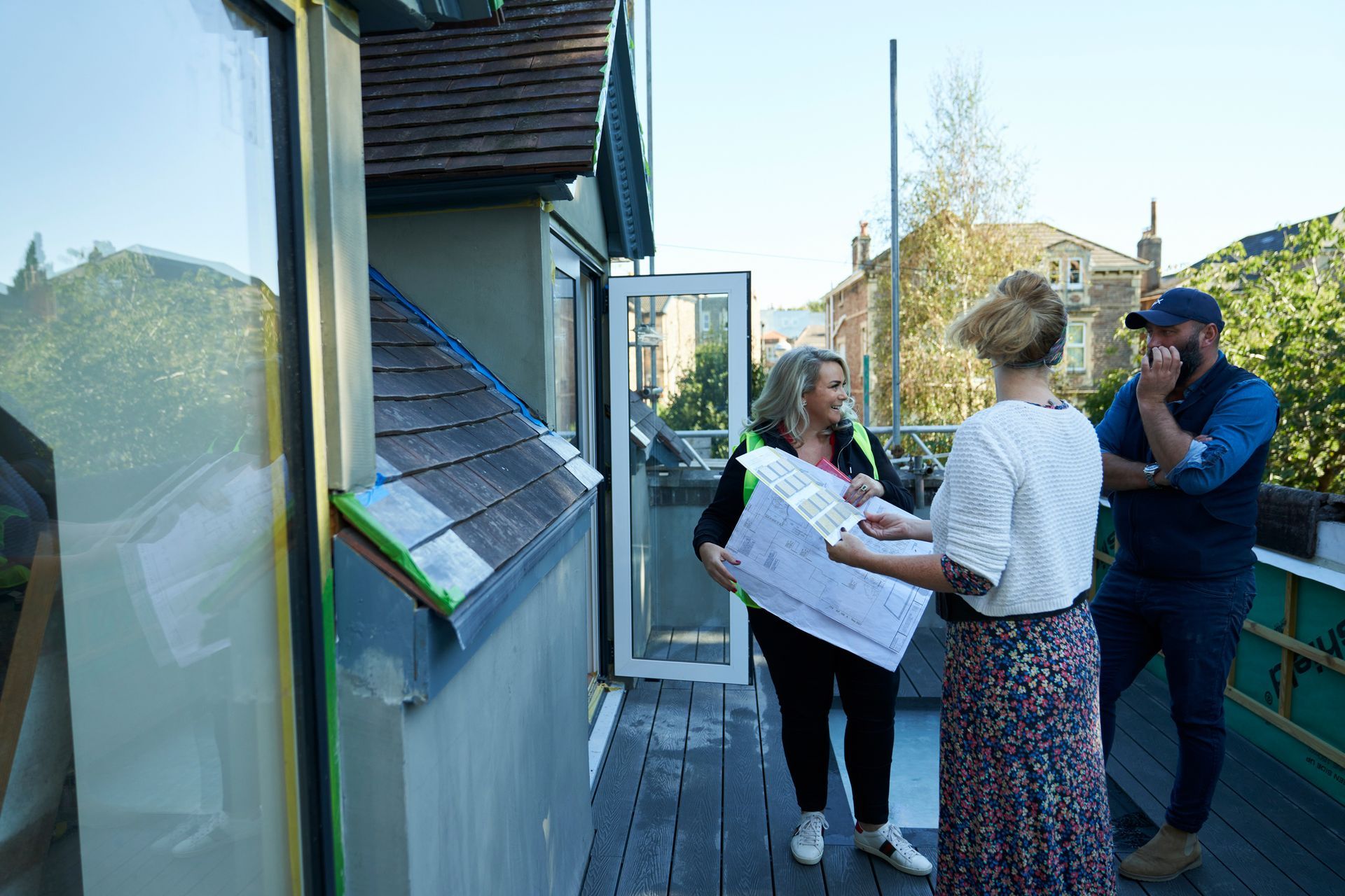Three people on a balcony reviewing blueprints. A woman holds plans; others look on with concerned expressions.