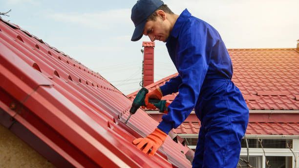 Roofer in blue jumpsuit and gloves, installing red metal roofing with a power drill on a sunny day.