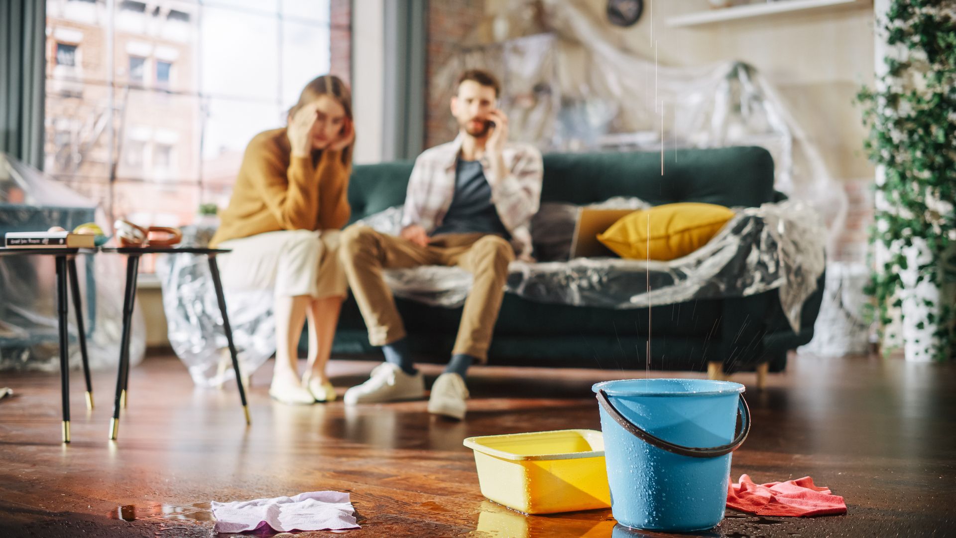 A couple distressed in a water-damaged living room. A bucket collects water, with plastic sheeting protecting furniture.