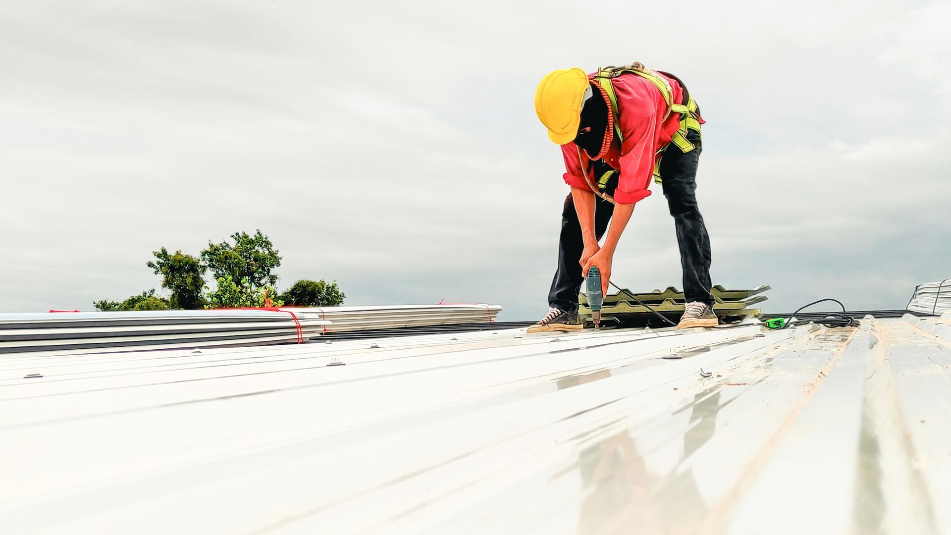 Roofer in safety harness using tool on a metal roof under an overcast sky.