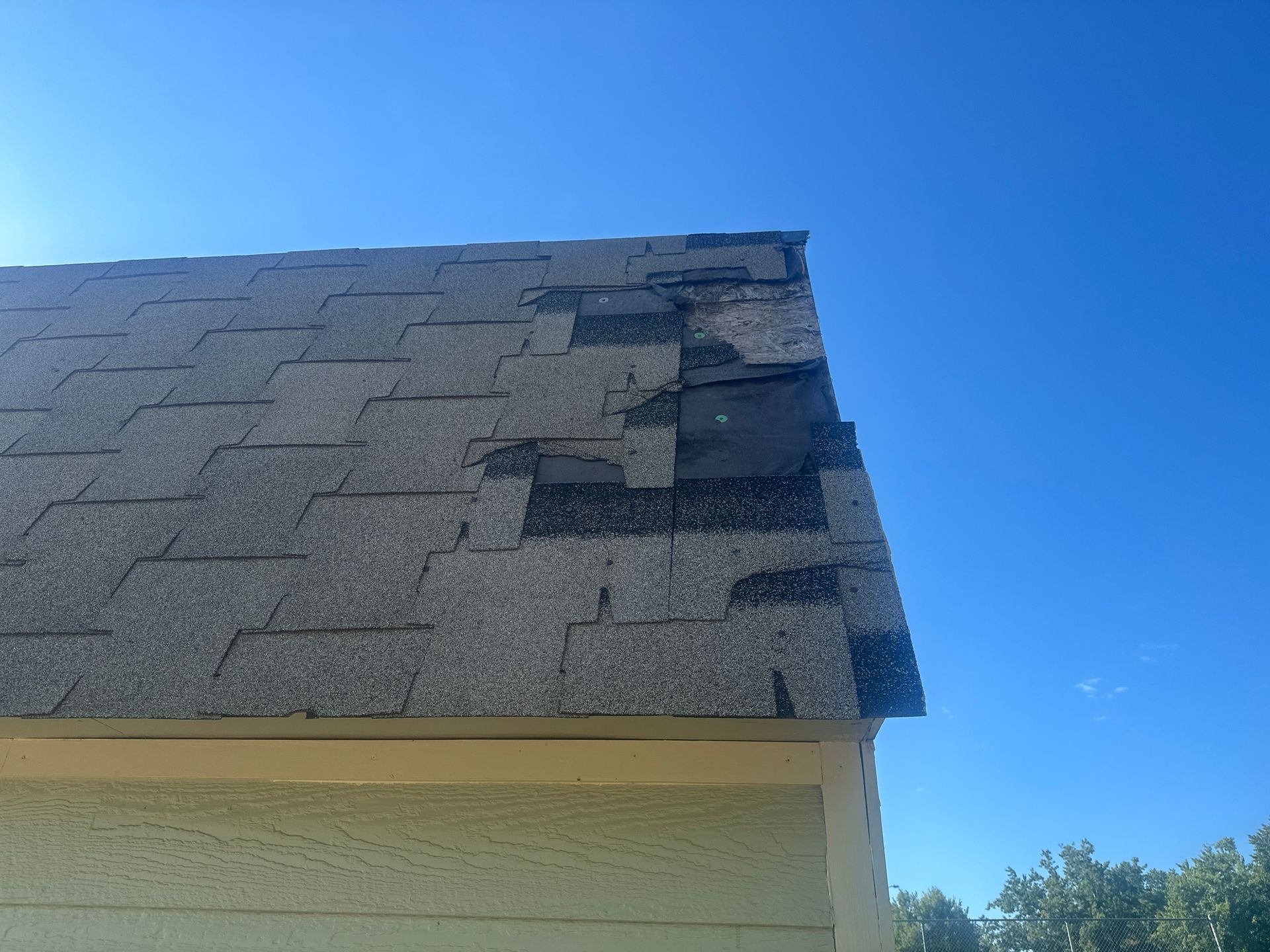 Damaged asphalt shingle roof against a bright blue sky. Torn and missing shingles visible.
