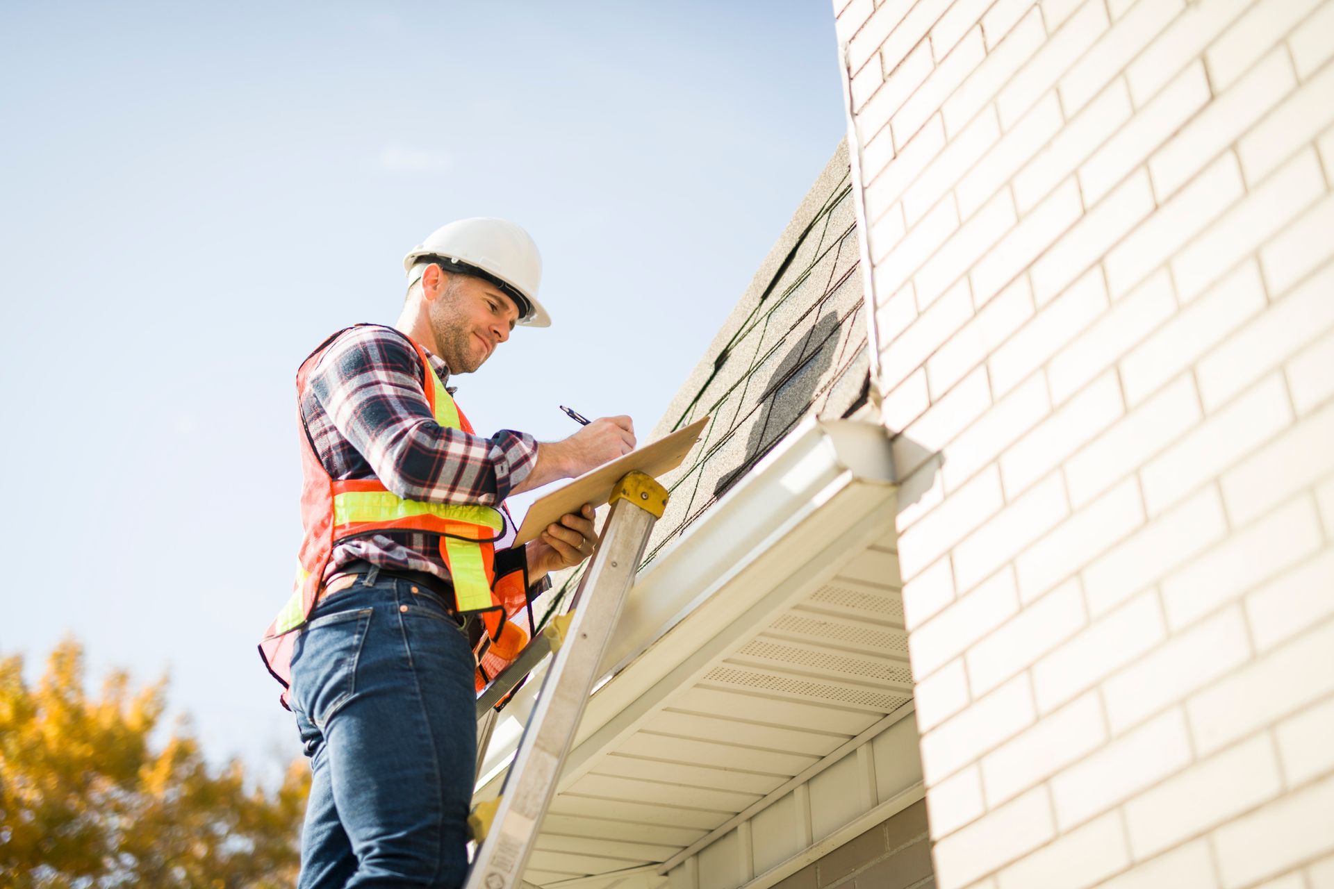 Man in safety gear on a ladder, inspecting a roof, taking notes on a clipboard.