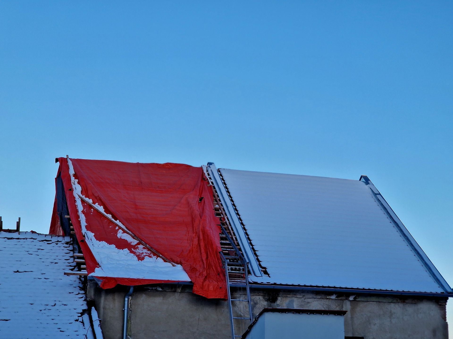 Snow-covered roof with a red tarp on one side, ladder against the building, clear blue sky.