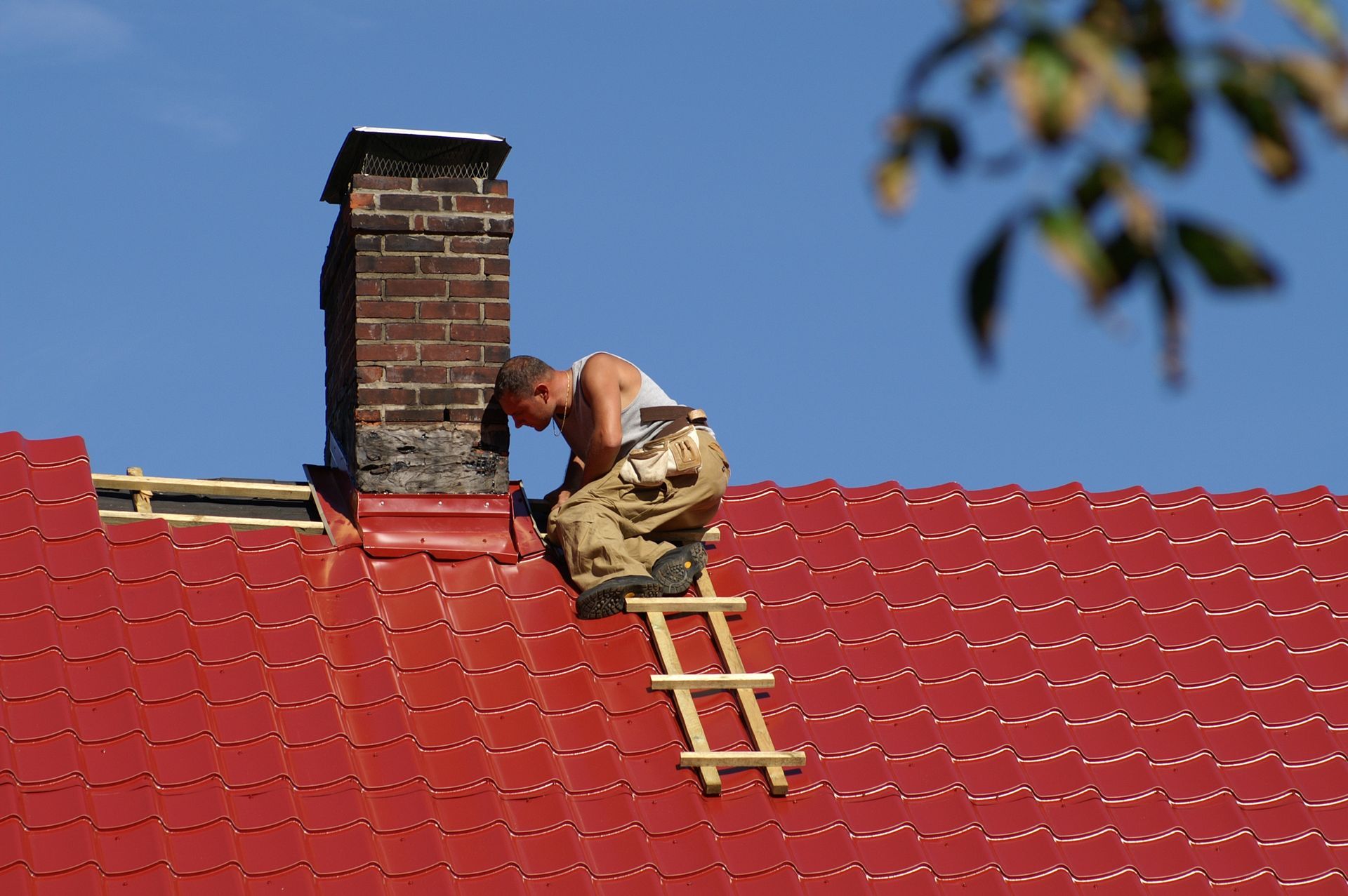 Roofer repairing a brick chimney on a red tiled roof under a blue sky.
