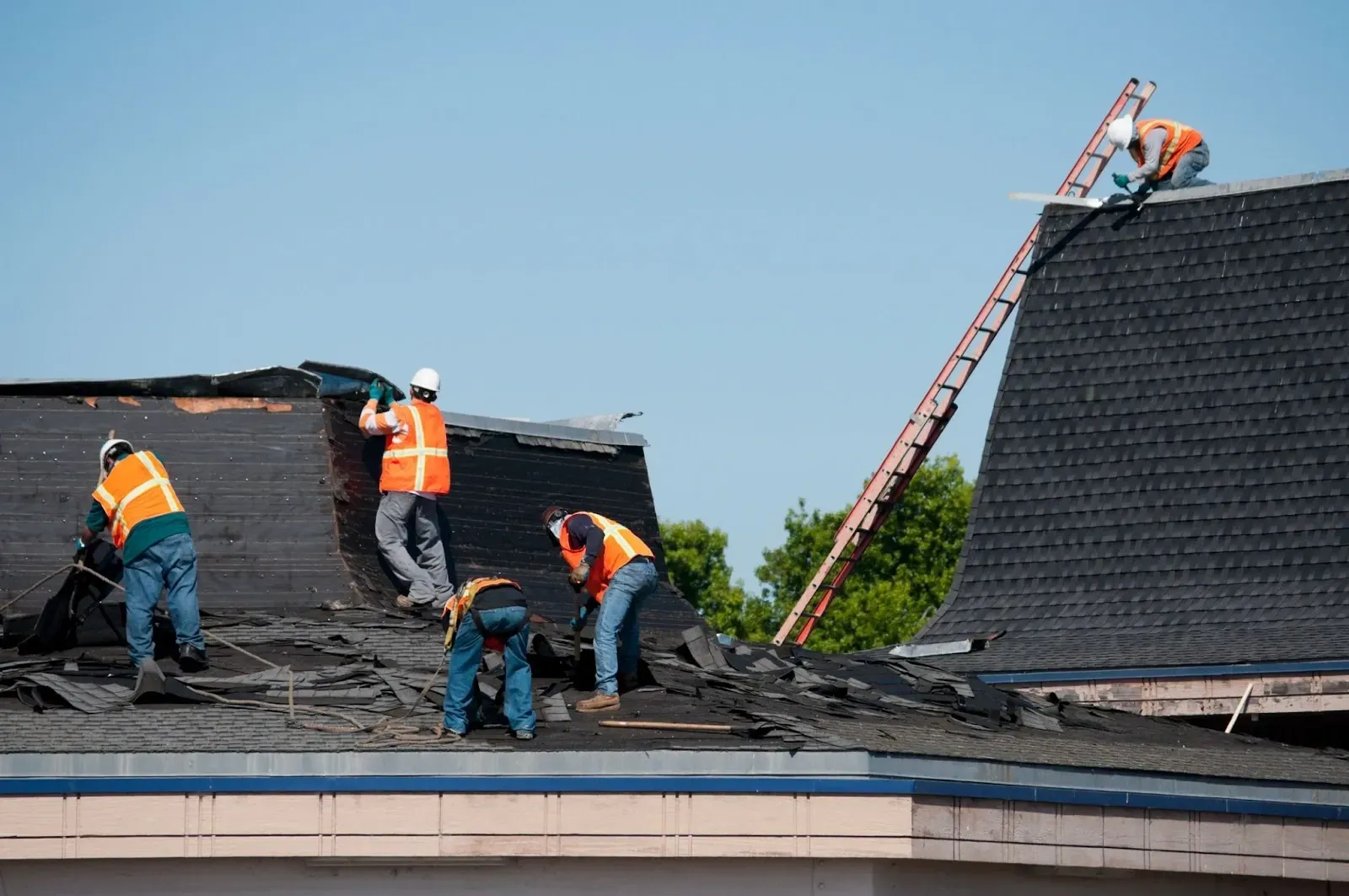 Roofers in orange vests and hard hats removing shingles from a dark roof on a sunny day.