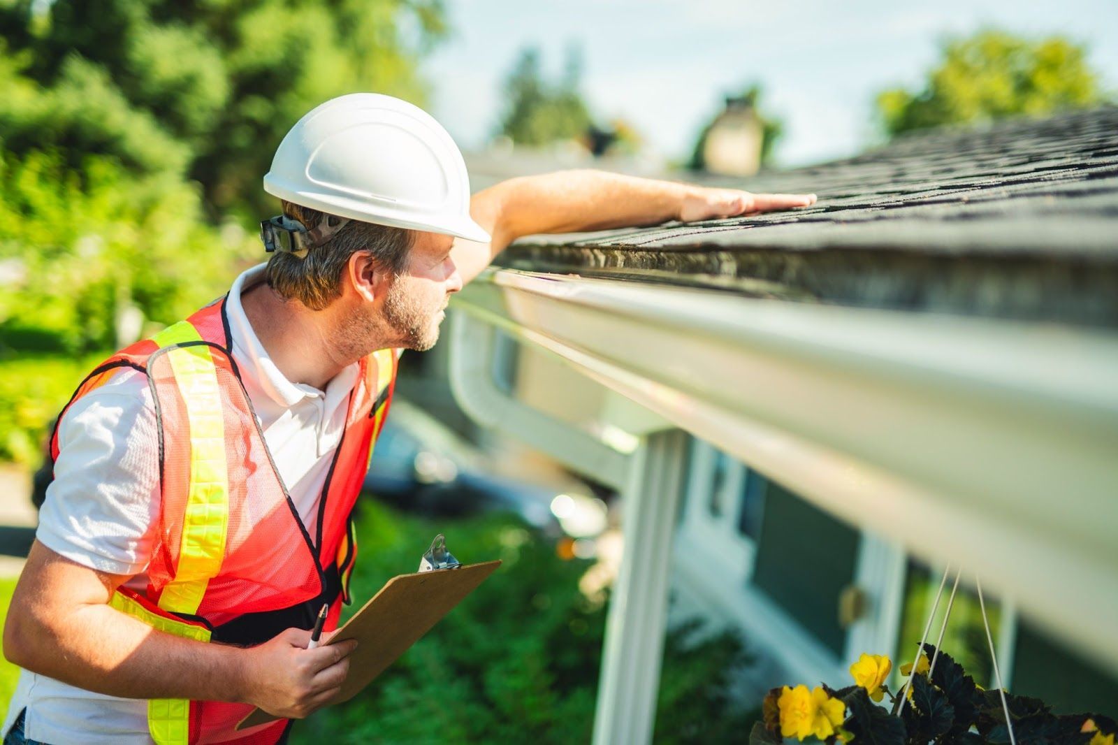Man in hard hat inspects roof gutter, holding clipboard. Outdoors, sunny.