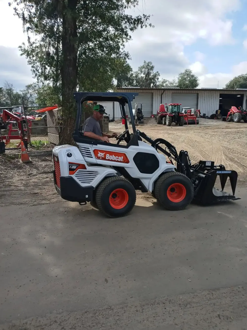 White skid-steer loader with red wheels and front bucket parked on a dirt lot near farm buildings
