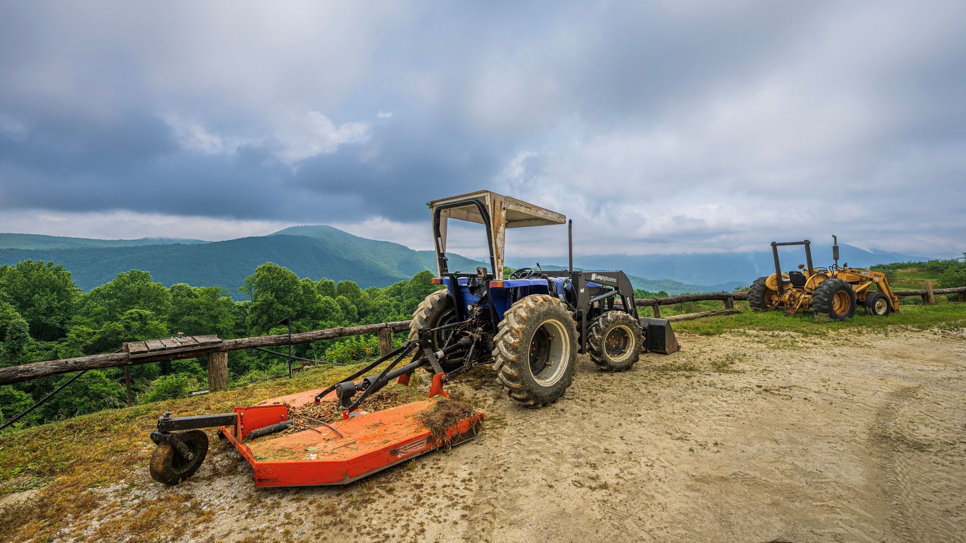 Blue tractor with mower attachment on a gravel road, with another tractor in a cloudy rural field.