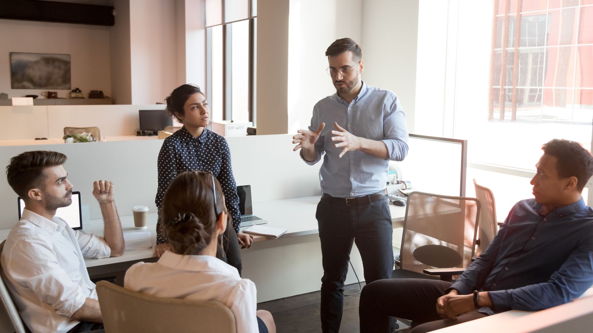 People in a meeting room listening as a man stands and presents with a whiteboard behind him.