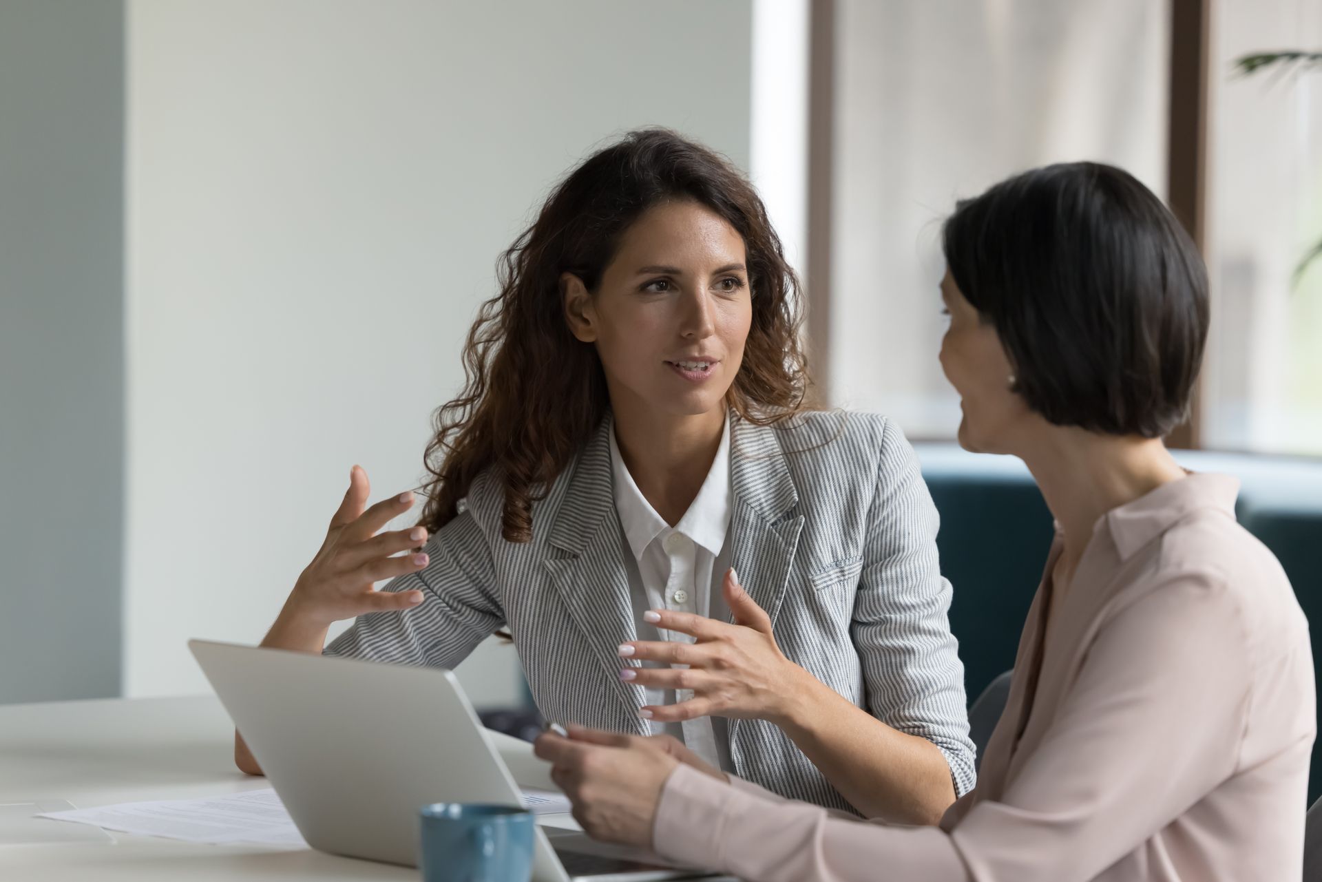 Two women talk at a table with a laptop in a bright office setting.