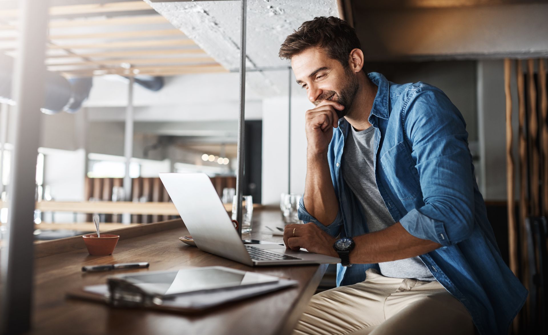 Man in a denim shirt using a laptop at a desk in a bright modern office