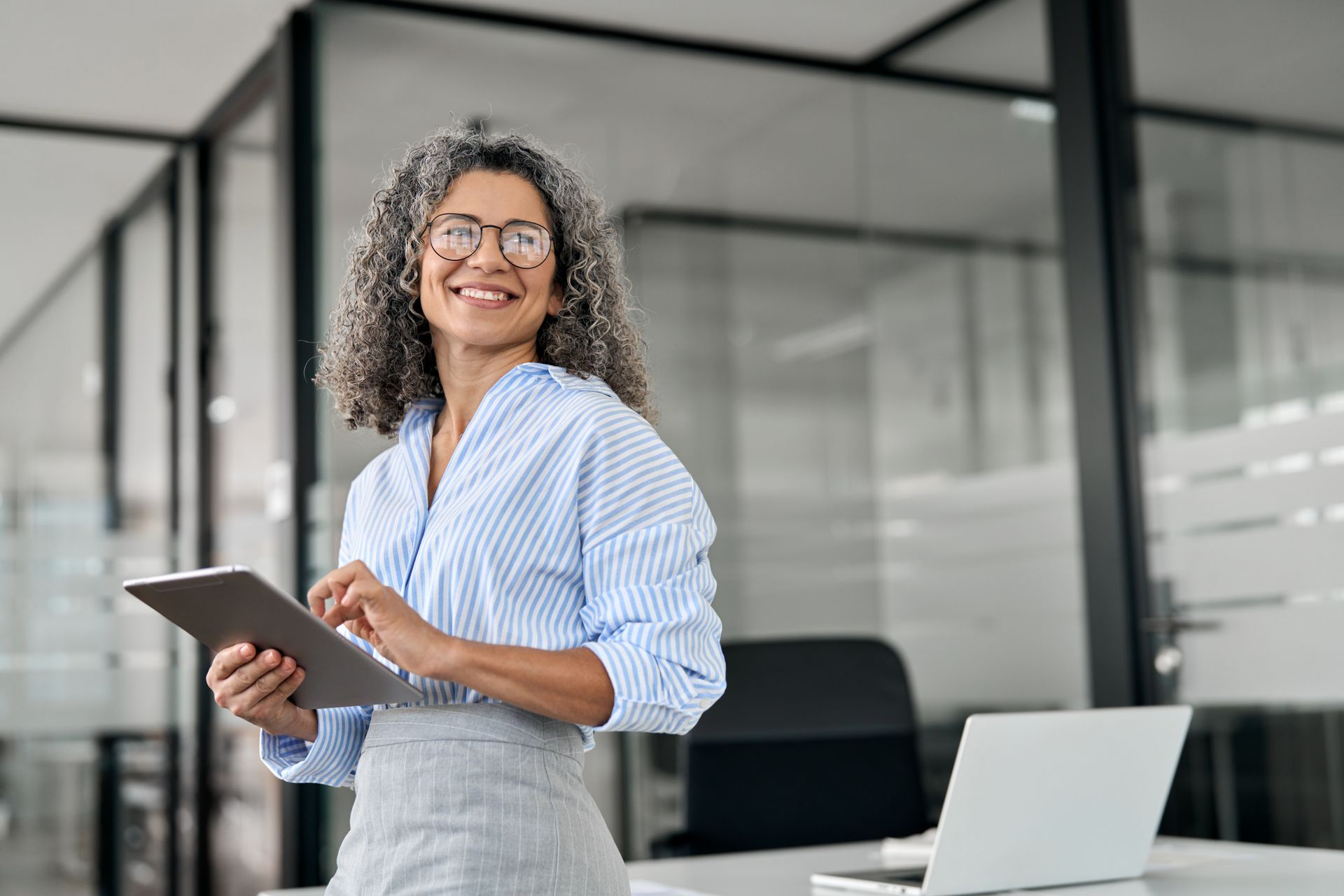 Smiling woman holding a tablet in a modern office with a laptop on the desk