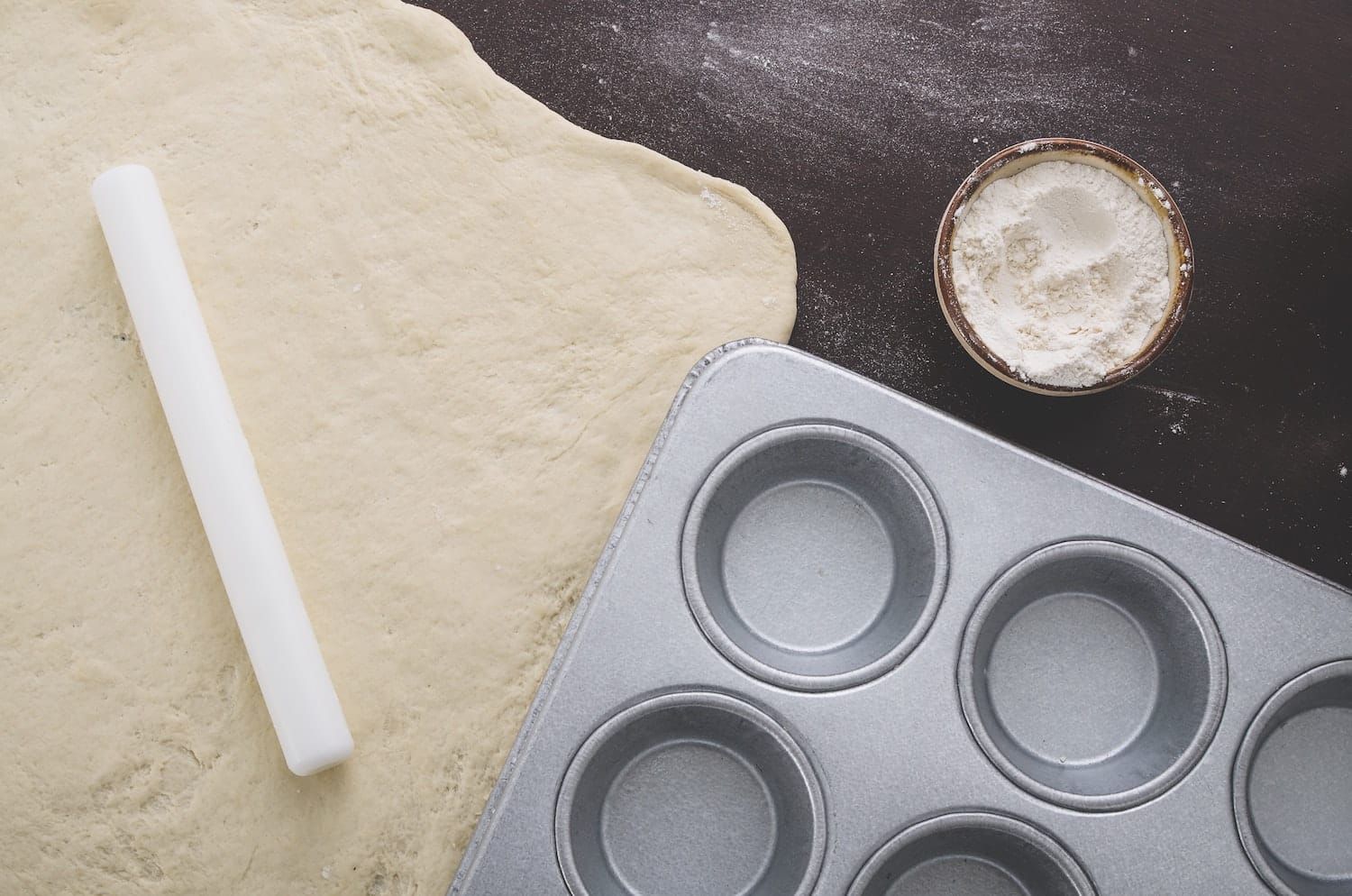 A rolling pin is sitting on a piece of dough next to a muffin pan.