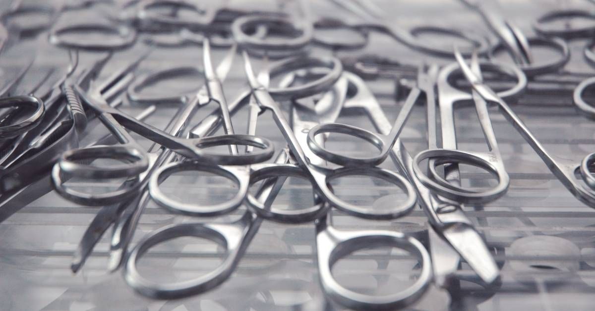 A pile of small medical scissors made of metal lying on a table. You can see their reflection in the