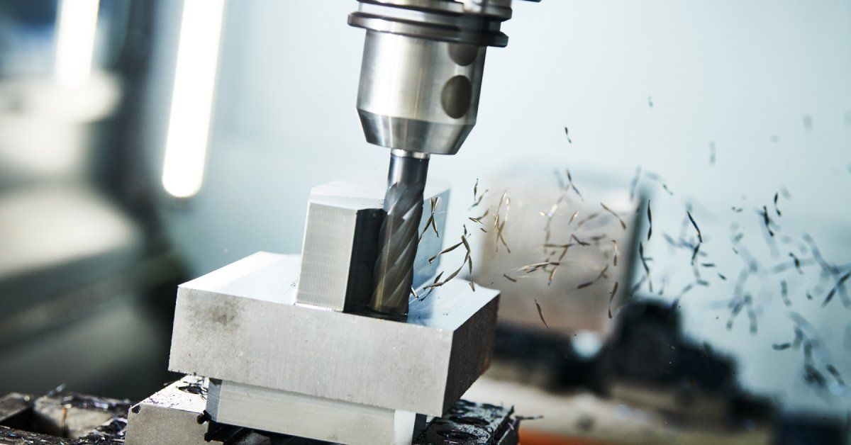 A CNC drilling machine boring holes into a block of silver metal with small shavings flying off.