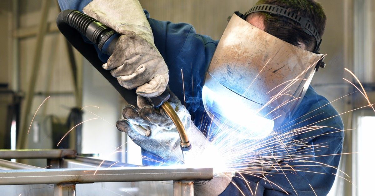 An industrial metalworker wearing a visor and gloves operates a welding torch with sparks coming off of the surface.