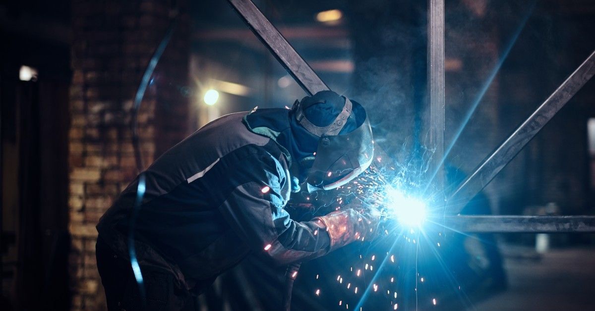 A man wearing protective clothing and a face mask uses a torch to fuse metal rods together in a factory.