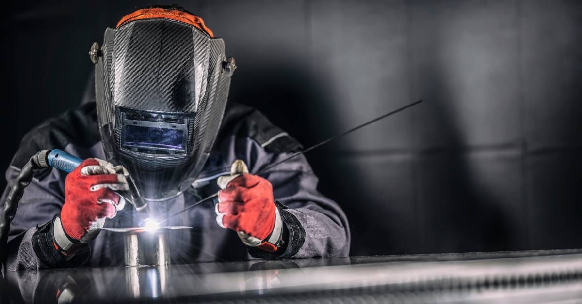 A worker wearing protective clothing and a face shield while using metal rods to create a weld on a small object.