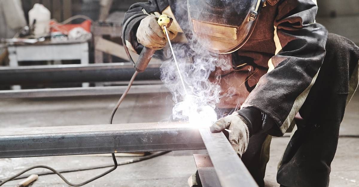 A worker kneeling in front of a metal structure and using a torch to make a weld between two of the structure's seams.