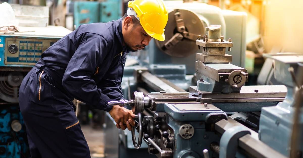 A worker in a blue jumpsuit and yellow hard hat operating a manual machining lathe on a factory floor.