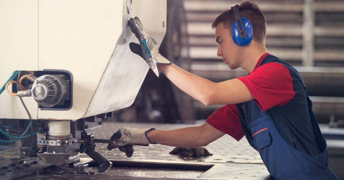 A factory worker wearing gloves and ear protection using a machine to press a piece of metal in his hand.