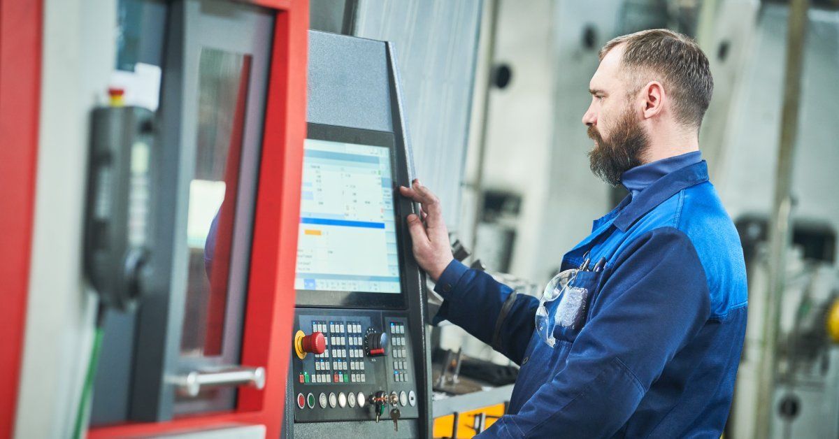 A man wearing a blue jumpsuit standing in front of a screen on a CNC machine entering commands for fabrication.