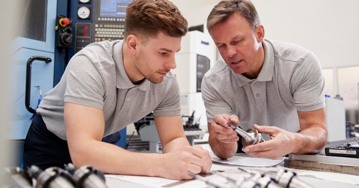 Two workers in gray shirts standing over a table with a number of manufactured components in front of a CAD machine.