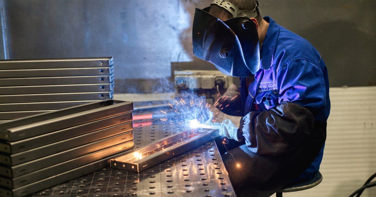 A worker wearing a face mask and protective gloves sitting at a bench using a torch to weld metal.