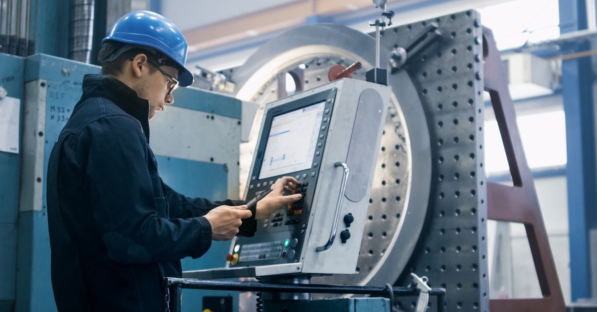 A worker wearing a black jacket and blue hard hat looking at a tablet computer while entering commands into a CNC lathe.