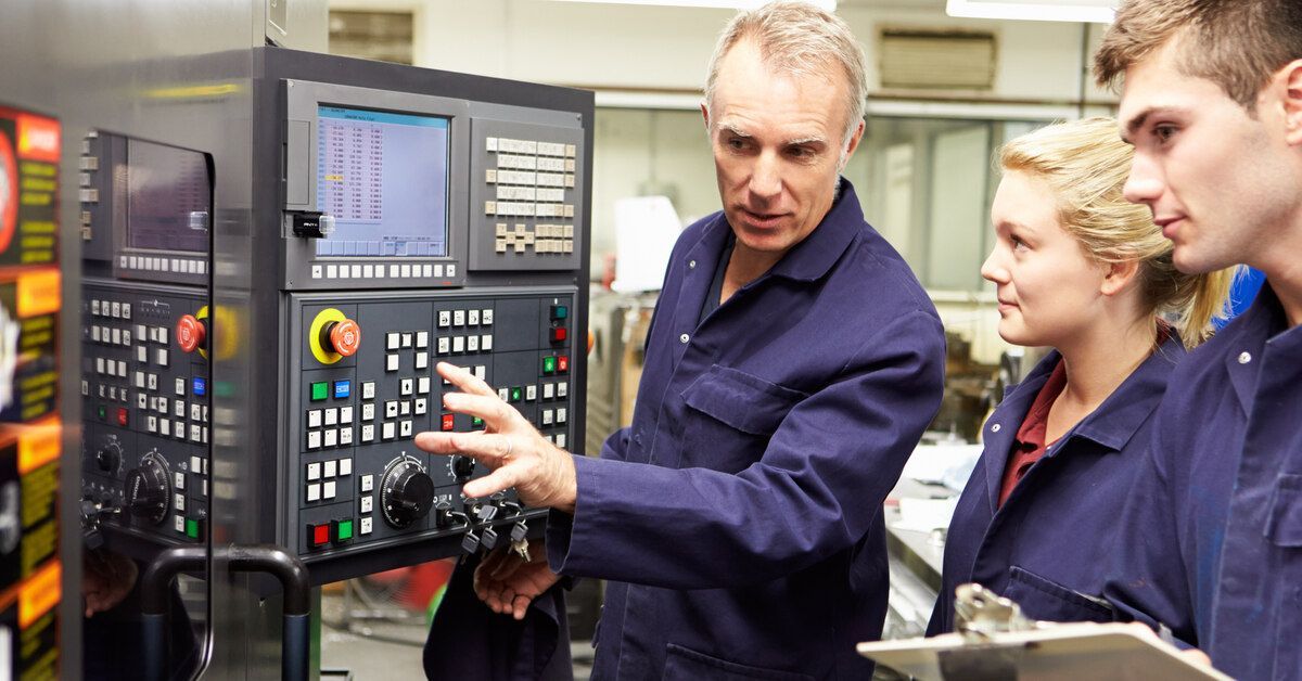 Three workers wearing blue jumpsuits standing in front of a CNC machine. One is showing the others how to use the equipment.