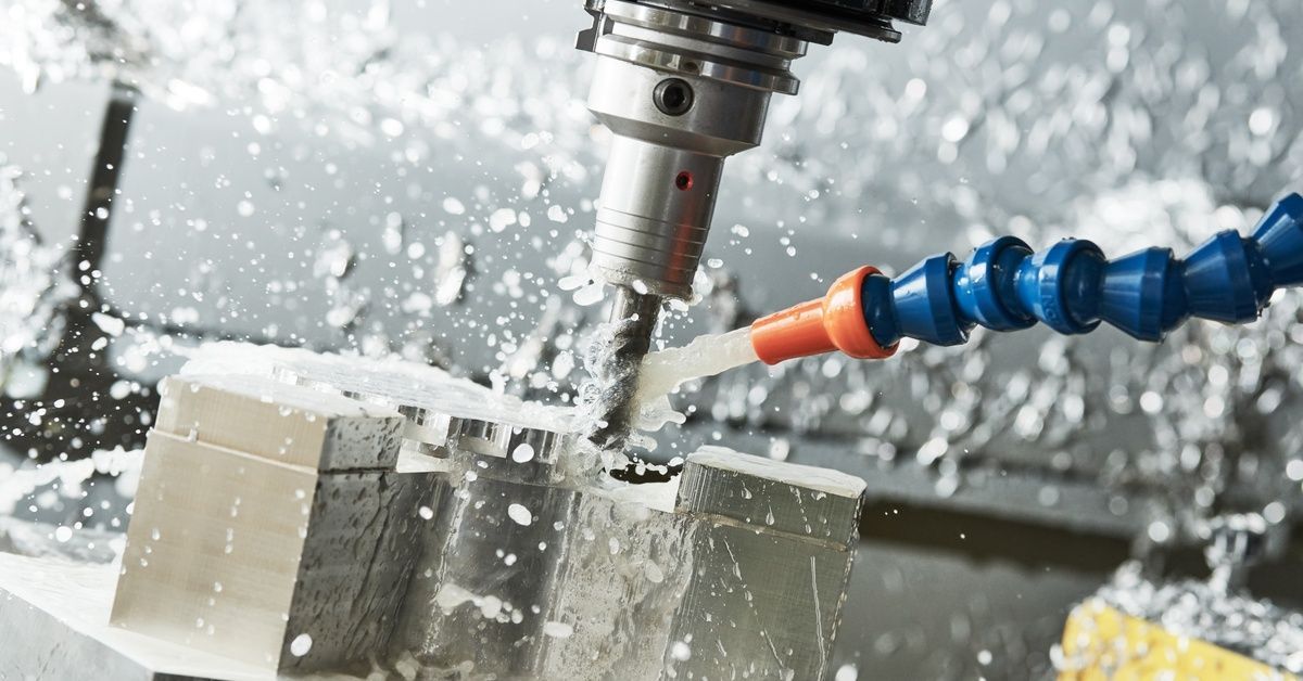 A close-up of a CNC machine with a drill bit removing material from a small metal piece, with a hose spraying liquid.