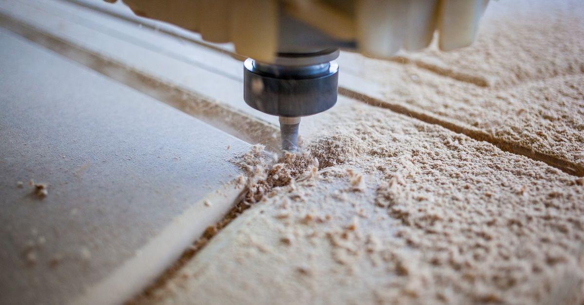 A close-up of a CNC machine removing material from a large piece of wood covered in sawdust with a drill bit.