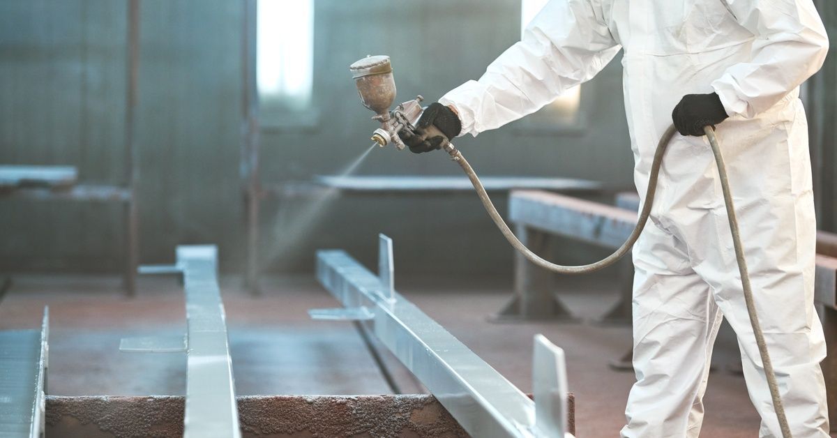 An industrial worker wearing a white protective jumpsuit uses a spray gun to apply a powder coating 