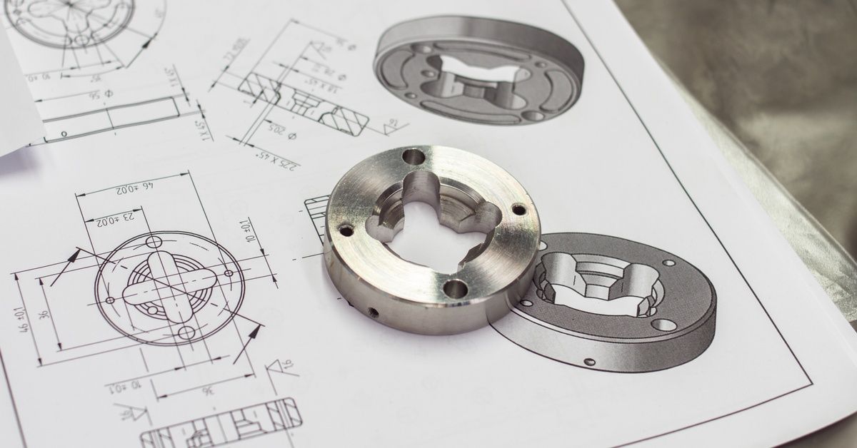 A circular metal component with small holes sits on top of a sheet of its printed design plans.