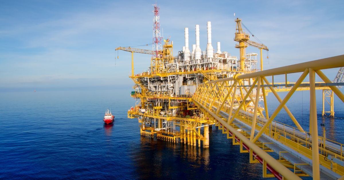 An offshore oil drilling platform in the middle of the ocean with multiple cranes and a red boat driving by.