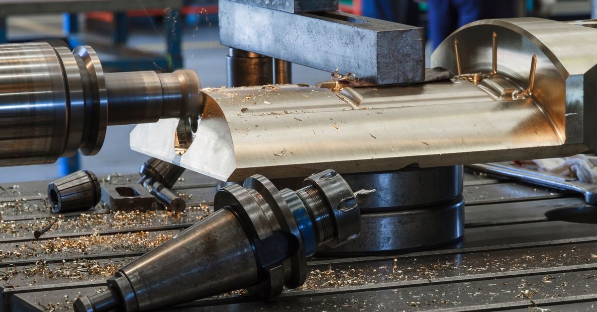 A CNC milling machine carves grooves into a semi-circular piece of bronze with a flat bottom on a large worktable.