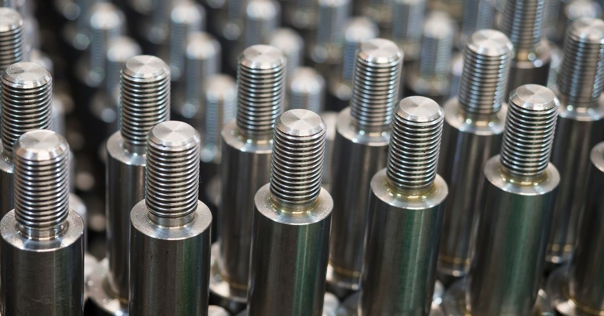 Close-up rows of steel metal workpieces with threaded grooves and a flat top. Metal pieces in the back are blurred.