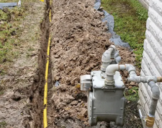 Gas meter and pipes beside a muddy trench along a house foundation