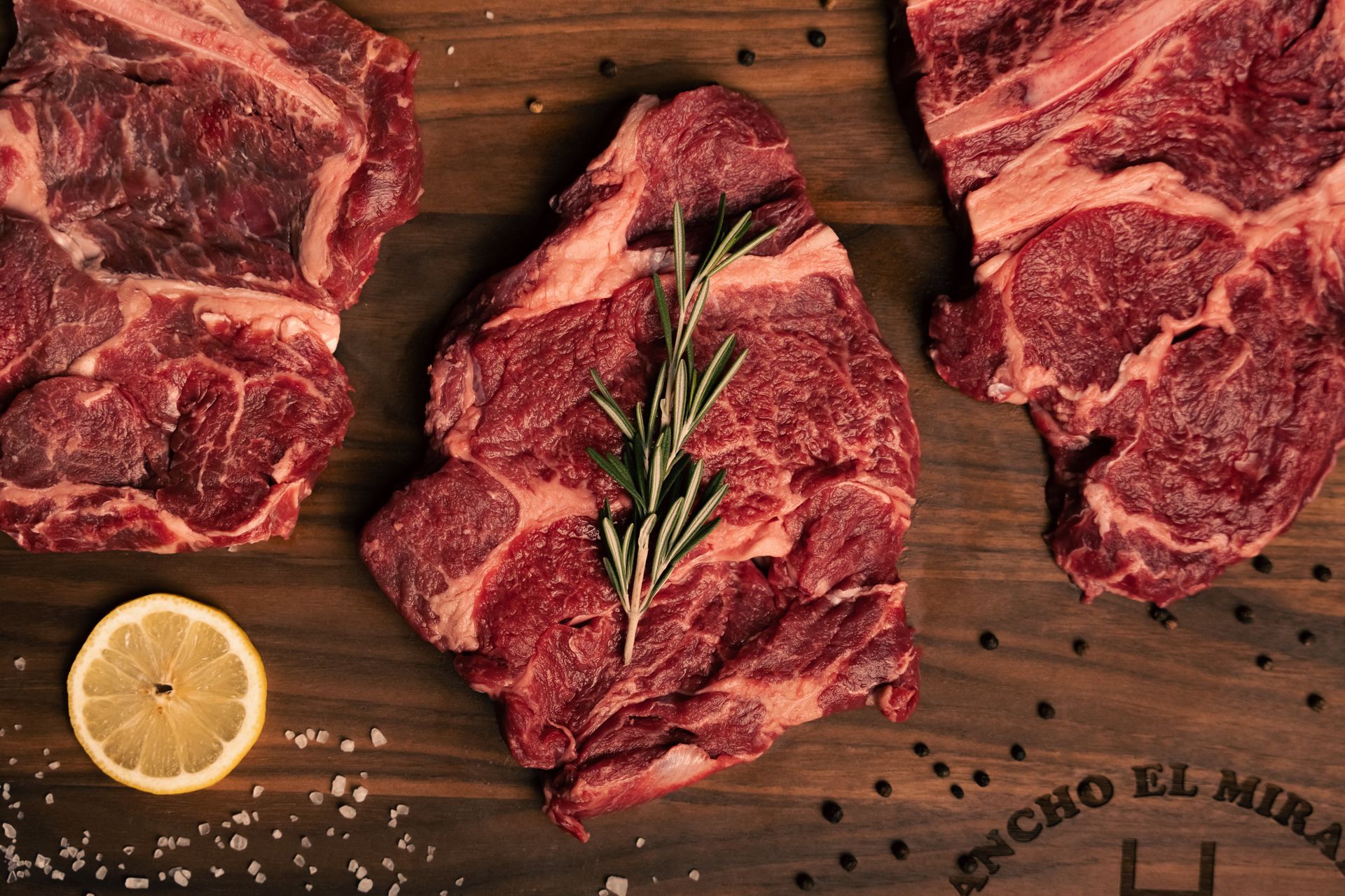 A piece of raw steak with rosemary on a wooden cutting board.