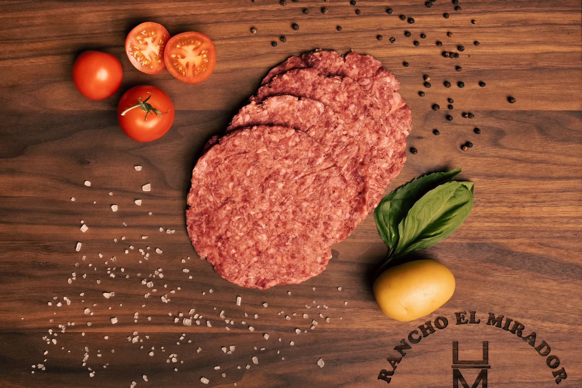 A wooden cutting board with hamburger patties and tomatoes on it.