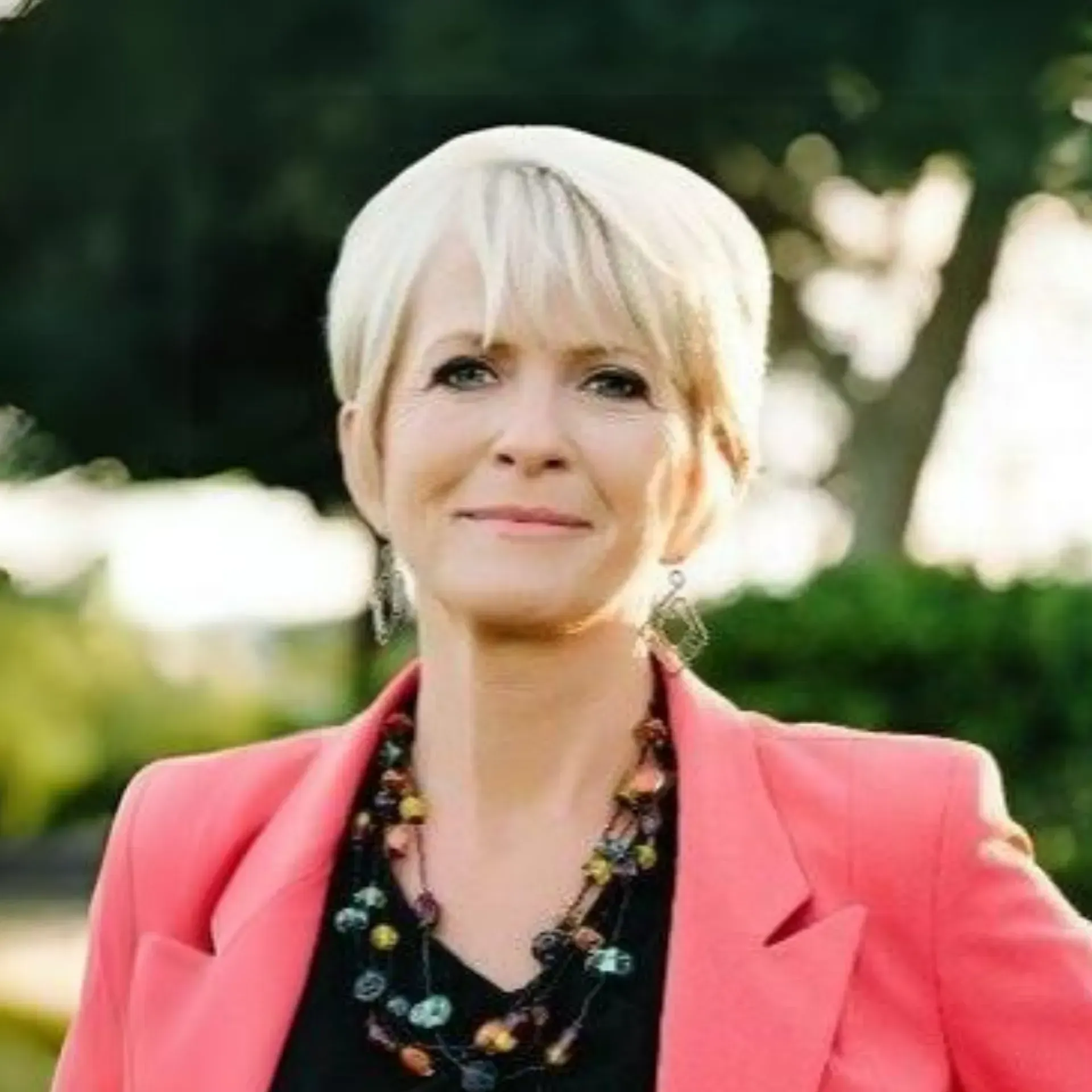 A woman wearing a pink jacket and a necklace is smiling for the camera.