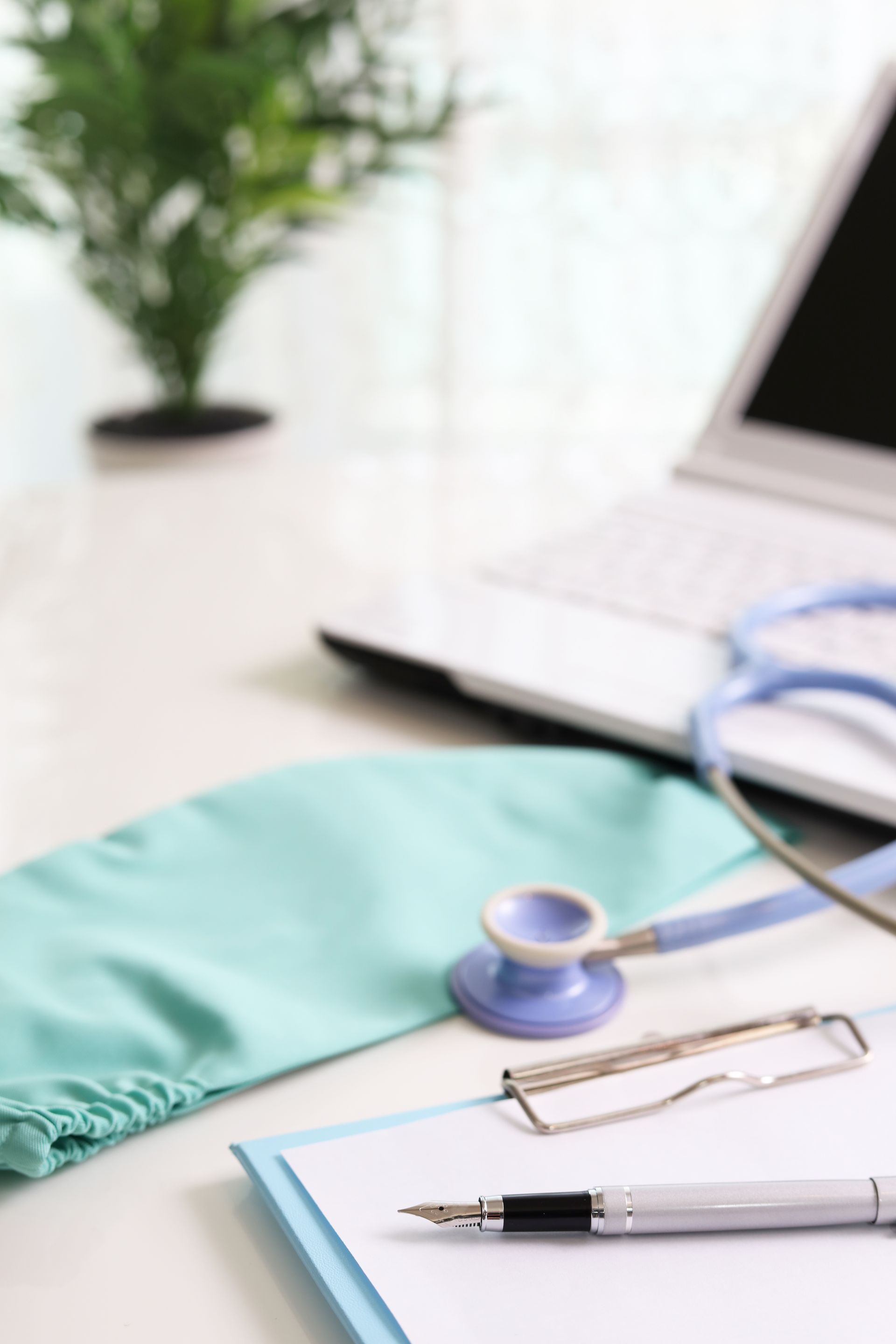 A doctor's desk: stethoscope, surgical cap, notepad with pen, laptop, and plant. A doctor's desk: stethoscope, surgical cap, notepad with pen, laptop, and plant.