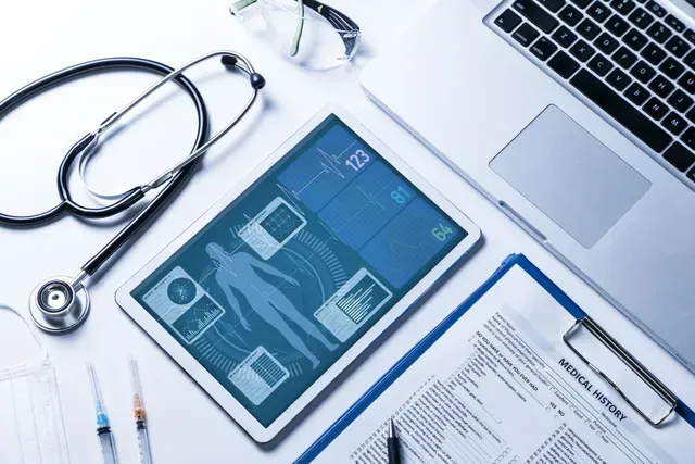 Medical devices on white surface: stethoscope, tablet with patient data, laptop, medical history form.