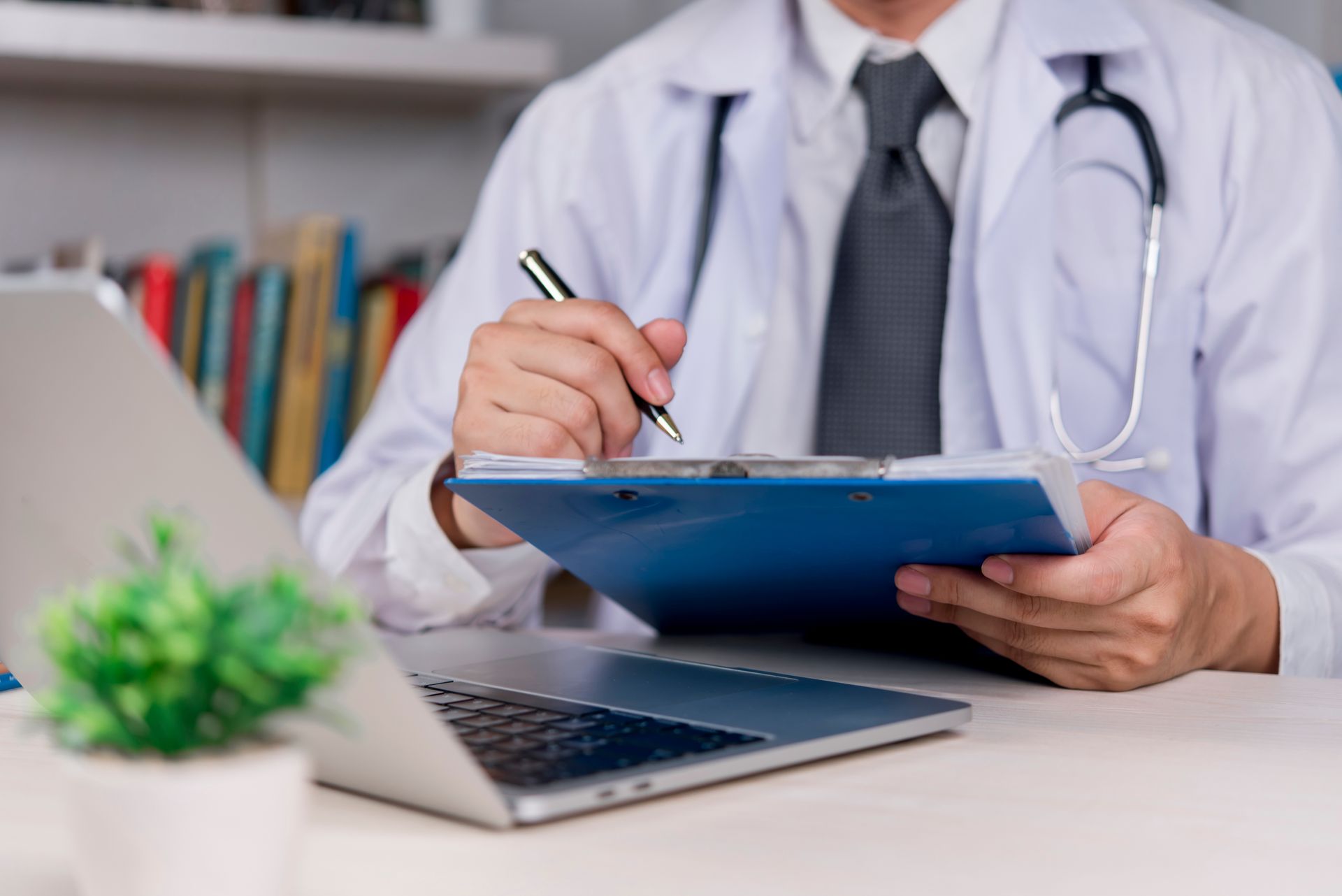 Doctor in white coat writing on a clipboard with a stethoscope, laptop, and plant on a desk Doctor in white coat writing on a clipboard with a stethoscope, laptop, and plant on a desk