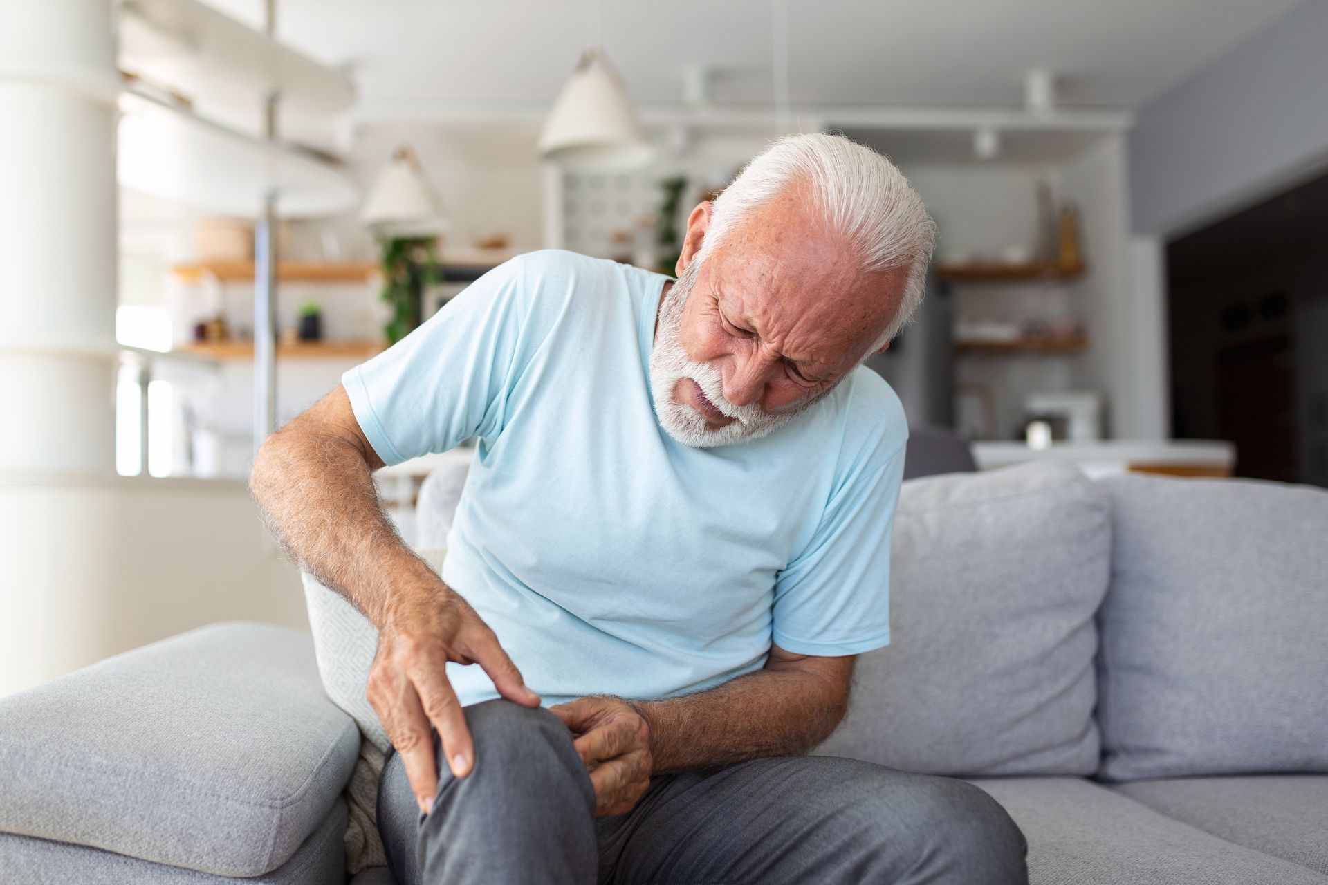 Older person on a sofa, grimacing and holding their knee in pain. Older person on a sofa, grimacing and holding their knee in pain.